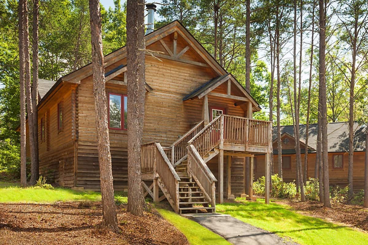 A rustic log cabin with a wooden staircase leading to a deck, surrounded by tall trees.