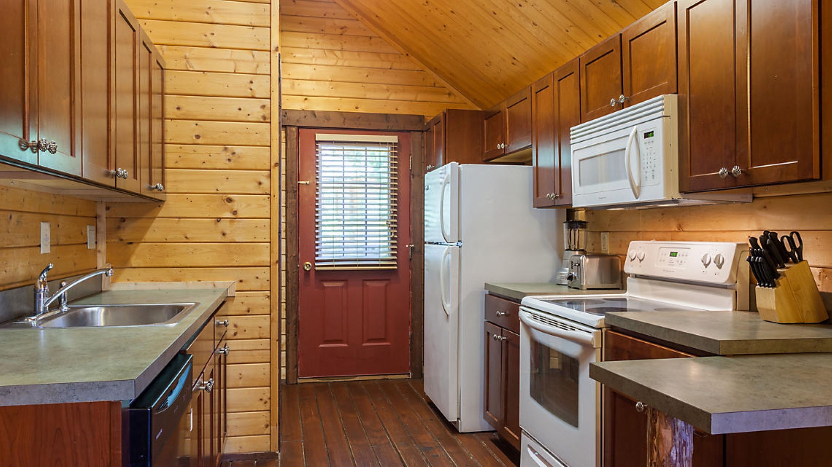 A compact kitchen with wooden walls, a refrigerator, a microwave, a stove, and a sink.