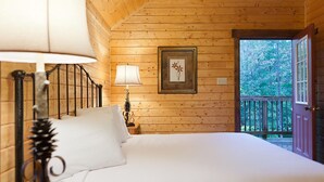 Un dormitorio de cabaña de madera con una cama, dos lámparas fijadas a la pared, un cuadro enmarco y una puerta que da a una terraza al aire libre.