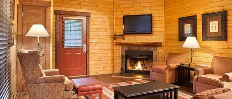A cozy log cabin interior with a fireplace, a television mounted above it, and a comfortable seating area with patterned chairs and a red bench.