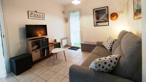 A living room with a sofa, a TV, a small table, and a chair. There are framed pictures on the walls and a window with blinds. - Maison 6 Pers 2 Chambres Bord de mer à Banastère (Sarzeau)