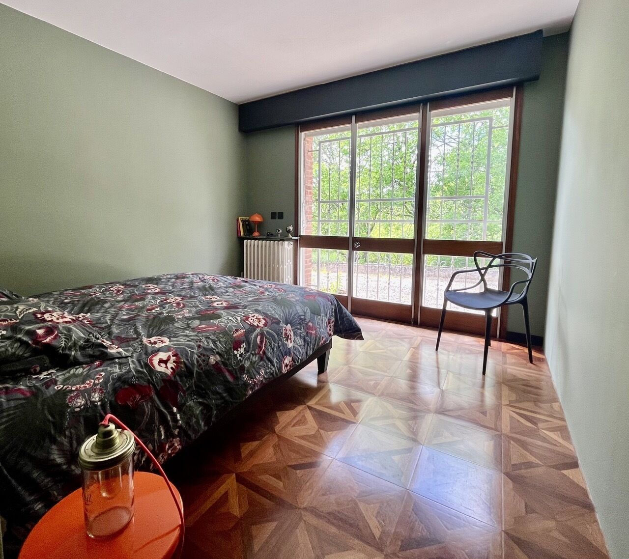A bedroom with a bed, a chair, a radiator, and a window with a view of greenery.