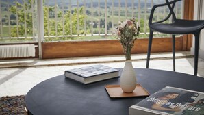 Una mesa redonda con un libro, un florero con flores y una revista en un balcón con vistas a zonas verdes y colinas.