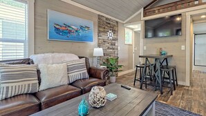 Una sala de estar acogedora con un sofá de cuero marrón, una mesa de centro con adornos, un cuadro de barcos, una pared de piedra y una zona de cocina con un ventanal grande.