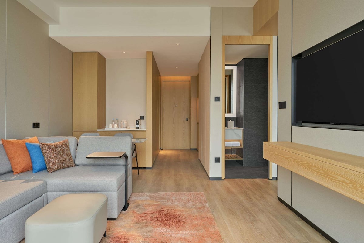 A modern hotel room with a sofa, ottoman, and a flat-screen TV mounted on the wall.