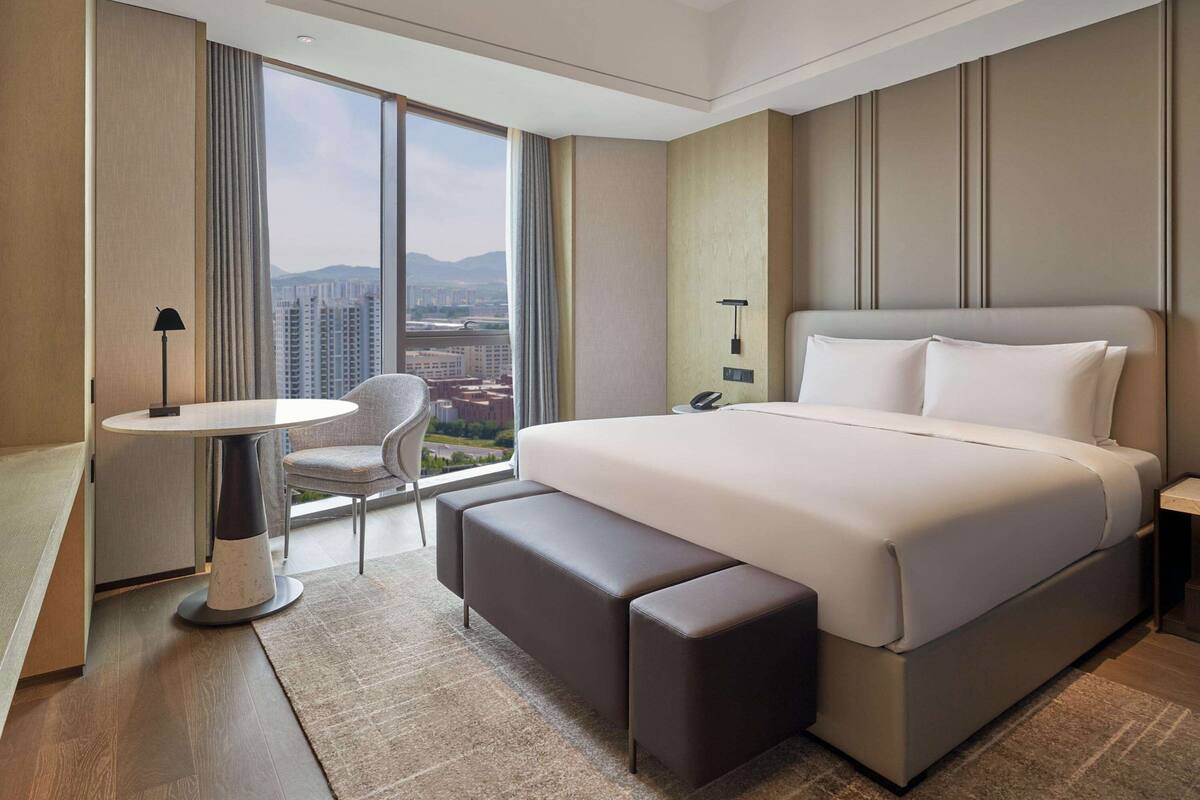 A modern hotel room with a large bed, a small round table, a chair, and a view of the cityscape through large windows.