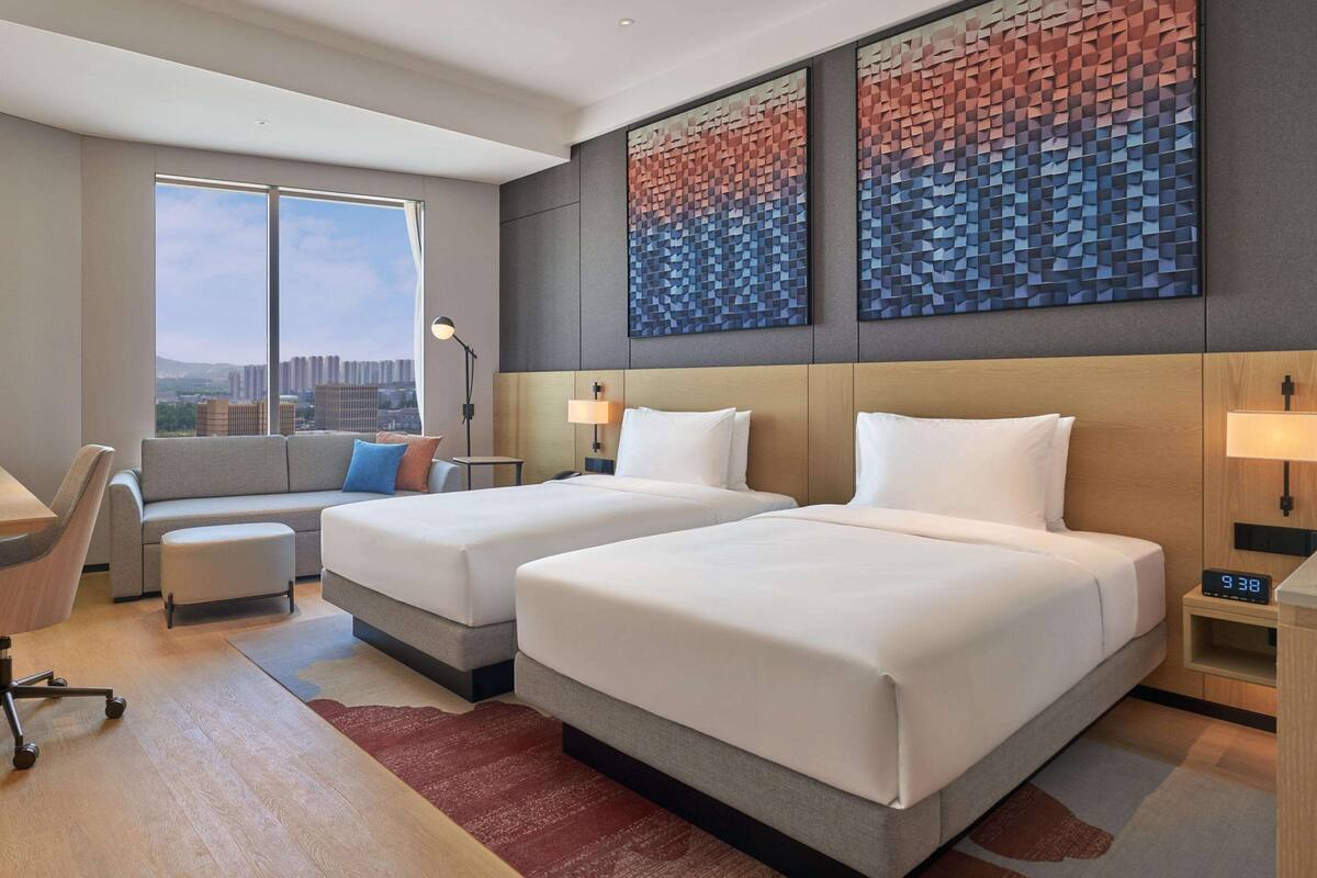 A hotel room with two beds, a sofa, a desk, and a large window offering a city view.