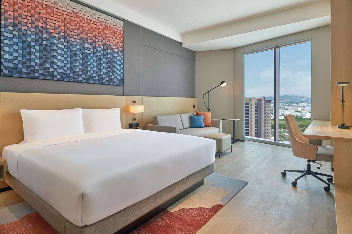 A modern hotel room with a large bed, a desk, a chair, and a view of the city through large windows.