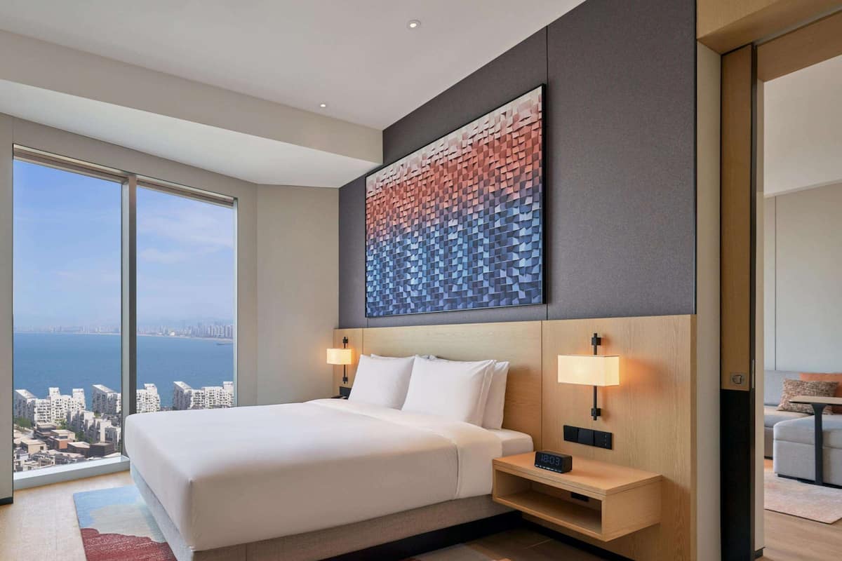 A modern hotel room with a large bed, two bedside lamps, a digital clock, and a cityscape view through large windows.
