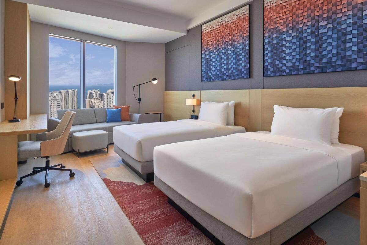 A hotel room with two beds, a desk, a chair, and a large window offering a city view.