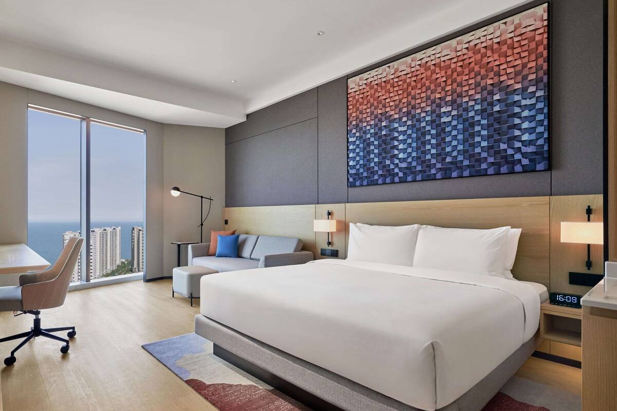 A modern hotel room with a large bed, a desk, and a chair. There is a view of the city through large windows.