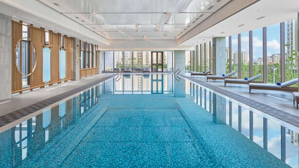 A modern indoor swimming pool with clear blue water, large windows, and lounge chairs along the sides.