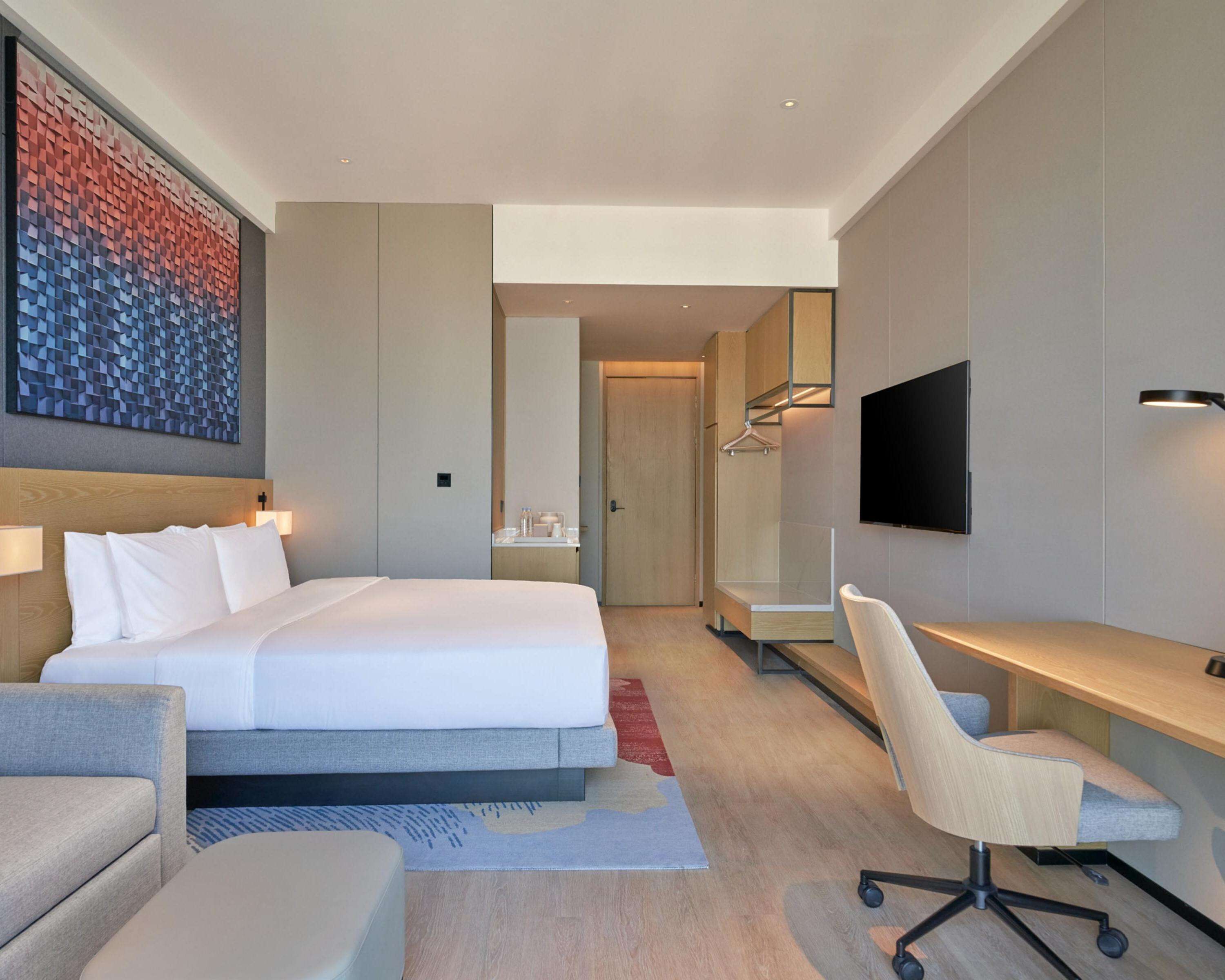 A modern hotel room with a large bed, a desk, a chair, a TV, and a wardrobe.