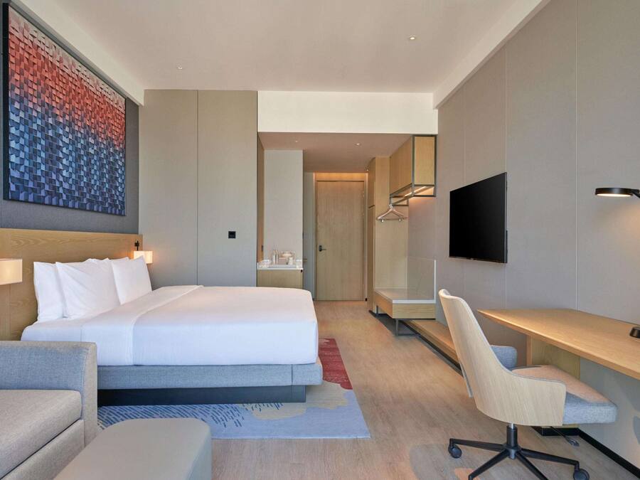 A modern hotel room with a large bed, a desk, a chair, a TV, and a wardrobe.