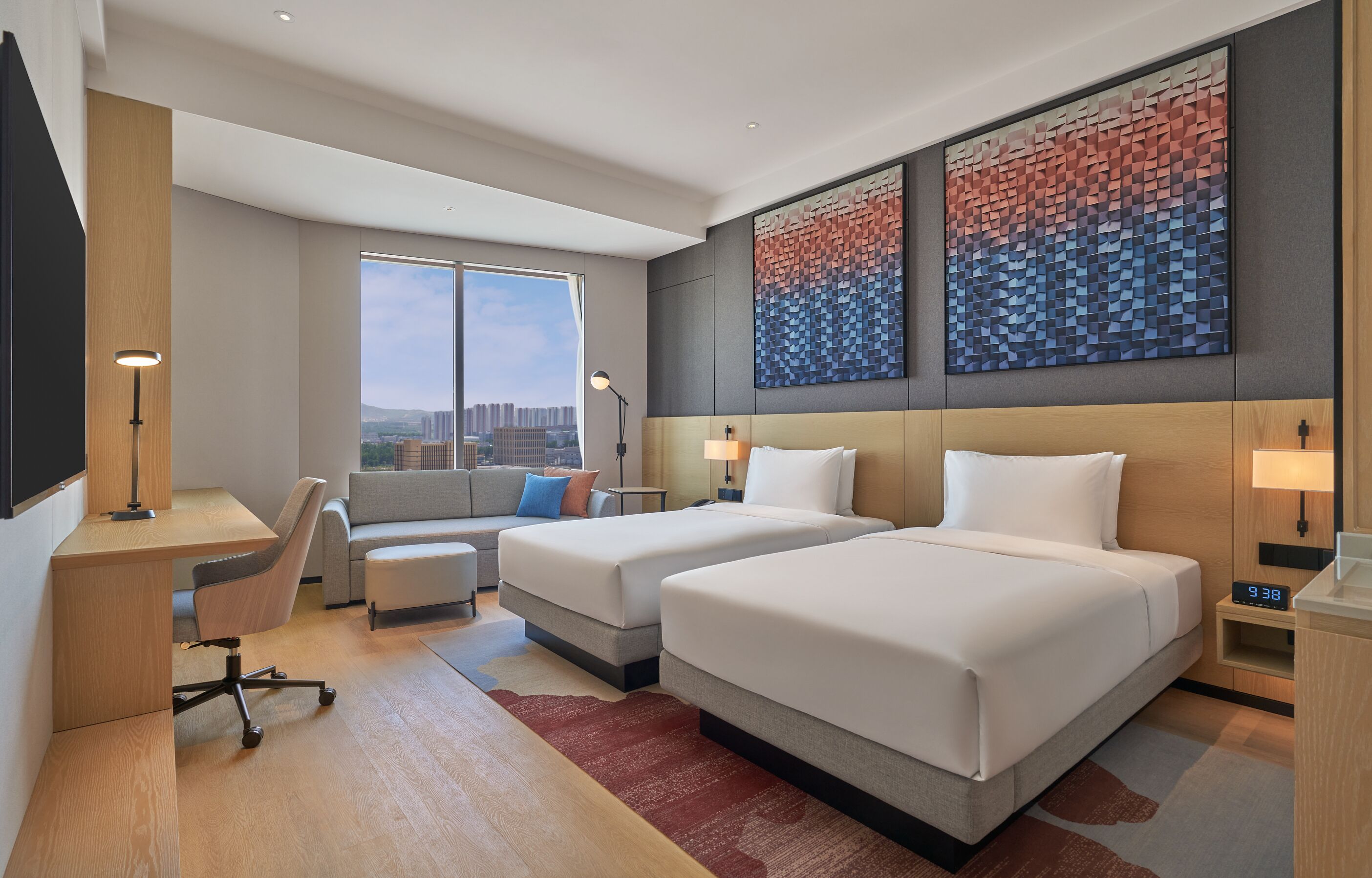 Quarto de hotel moderno com duas camas, uma escrivaninha, um sofá e uma janela grande com vista para a cidade.