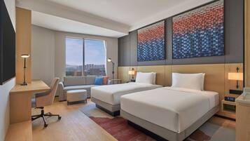 Quarto de hotel moderno com duas camas, uma escrivaninha, um sofá e uma janela grande com vista para a cidade.