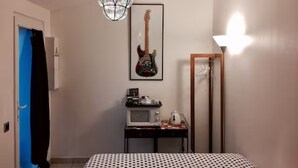 Un dormitorio con cama, un lámpara colgante, una guitarra en la pared, una mesita con una tetera y un perchero.