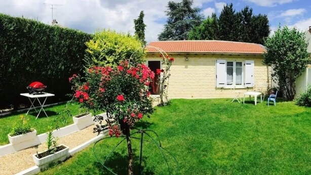 Una casa con tejado de tejas rojas, un pequeño jardín con un árbol y un parterre de flores, y una silla blanca en el exterior.
