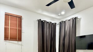 Ein Zimmer mit einem gelben Bett, dunklen Vorhängen, einem Deckenventilator und einem an der Wand montierten Fernseher.