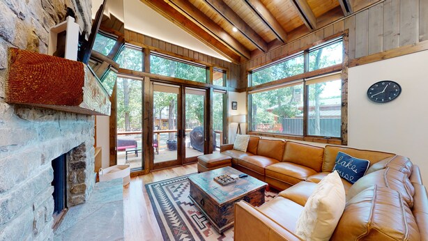A cozy living room with a stone fireplace, leather sofa, and large windows offering a view of the outdoors. - Private Hot tub, 1mi to Shangri La, next to Battlefield Par 3, & Walk to Lake (Afton)