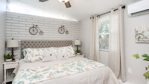 A bedroom with a white brick wall, two wall-mounted bicycles, a tufted headboard, a bed with a floral bedspread, two bedside tables with lamps, and a window with curtains. - Comfortable , newly remodeled, close to Siesta Key (Sarasota)
