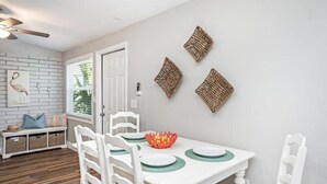 A dining area with a white table, green placemats, and a decorative bowl. There are white chairs and a bench with baskets. The walls have a white brick pattern and three woven wall hangings. - Comfortable , newly remodeled, close to Siesta Key (Sarasota)
