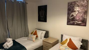 A twin bedroom with two beds, a nightstand, a lamp, a painting of a Buddha, and a window with curtains.