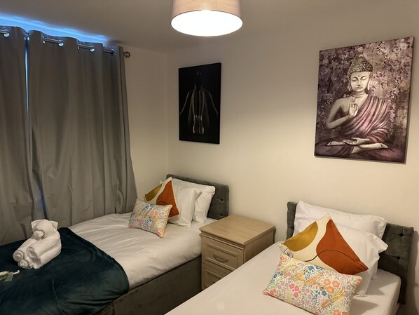 A twin bedroom with two beds, a nightstand, a lamp, a painting of a Buddha, and a window with curtains. - Luxurious 2-bed Apartment in Maidstone Kent (Maidstone)