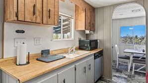 A compact kitchen with wooden cabinets, a microwave, a sink, and a window overlooking a scenic view. - Cosy Desert Hideaway (Tucson)