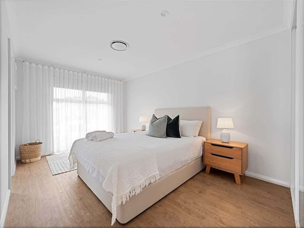 A bedroom with a large bed, a bedside table with a lamp, a window with sheer curtains, and a wooden floor.