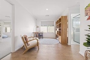 A spacious room with a wooden floor, a white wall, a large window, a desk, and a bookshelf.