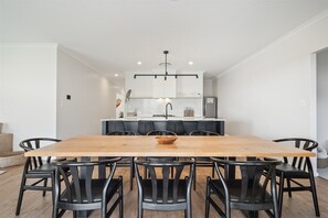 Un espace repas moderne avec une table en bois et des chaises noires, une cuisine en arrière-plan et un luminaire suspendu au-dessus.