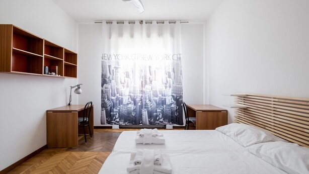 Una camera d'albergo moderna con una scrivania in legno, una sedia, un letto con asciugamani ordinati e vista sulla città dalla finestra.