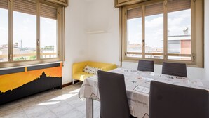 Zona pranzo con tavolo apparecchiato per quattro, ampie finestre con tende, un divano giallo e vista sulla città.