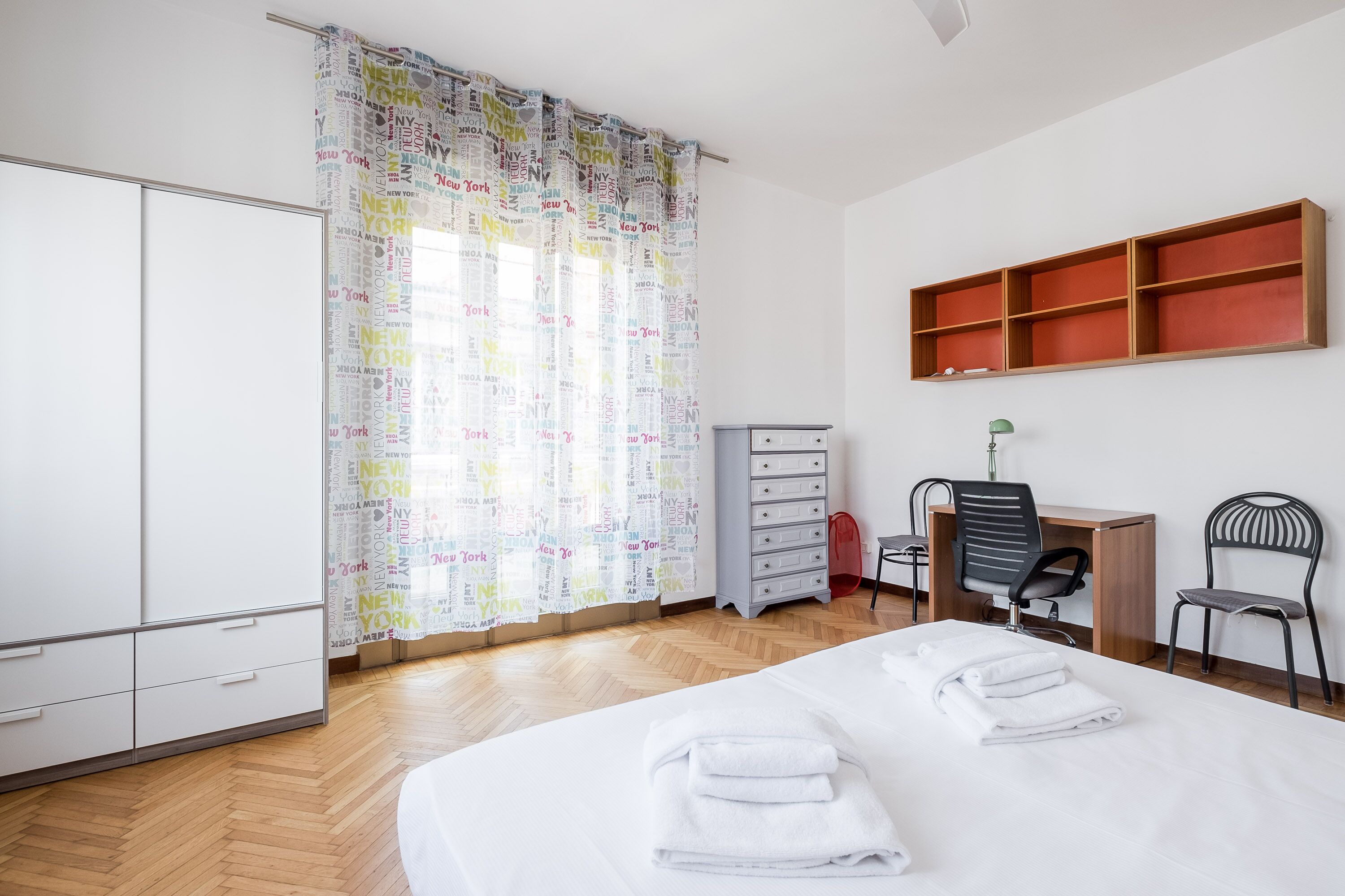 A hotel room with a bed, a desk, a chair, a wardrobe, and a shelf.