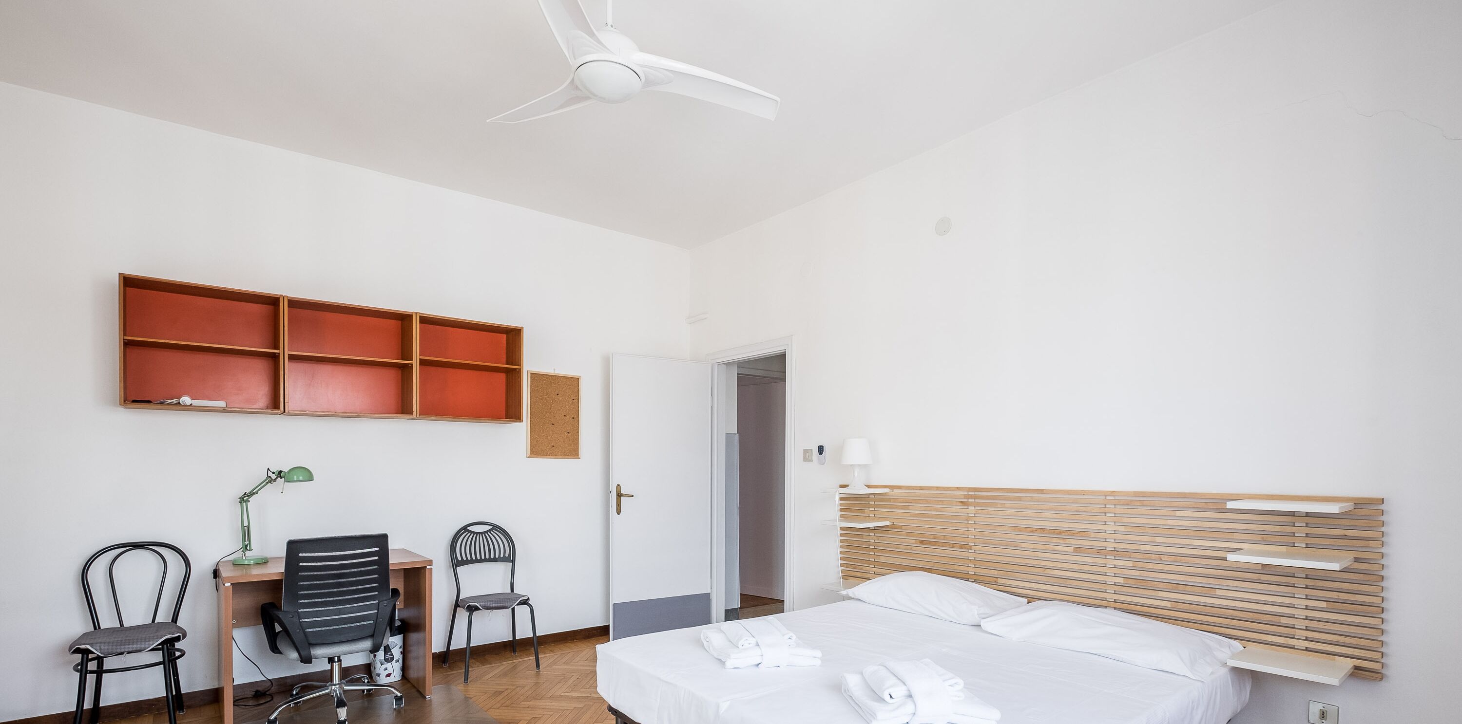 Een hotelkamer met een bed, een bureau met stoel, een plafondventilator en een wandplank.