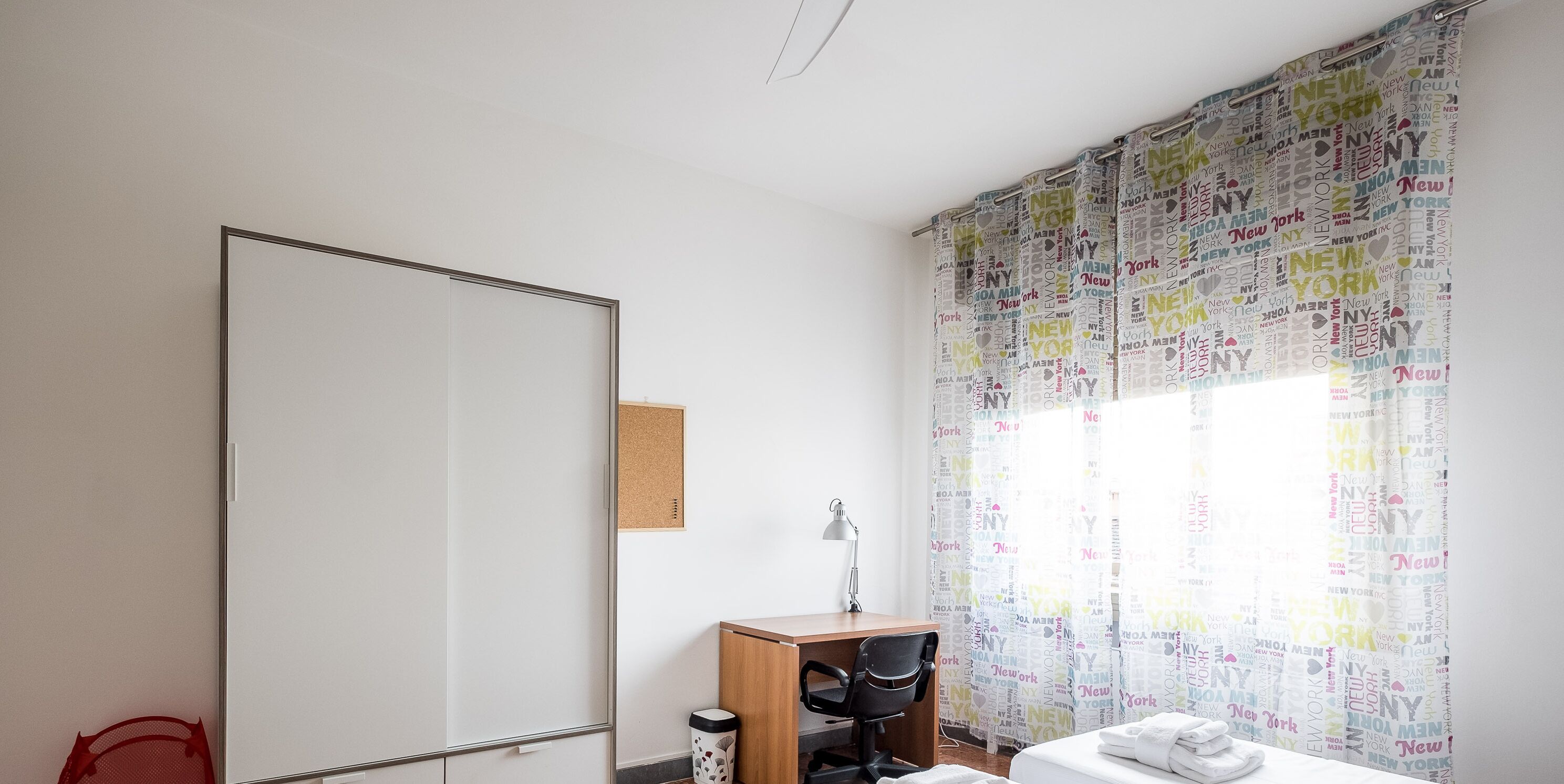 Une chambre à coucher comprenant un lit, un bureau, une chaise, une chaise rouge, une poubelle et un rideau orné de noms de villes.