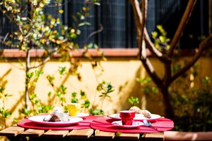 Ein Holztisch mit roten Tischsets, Tellern mit Speisen und Getränken, und eine gelbe Wand mit Grünpflanzen im Hintergrund.