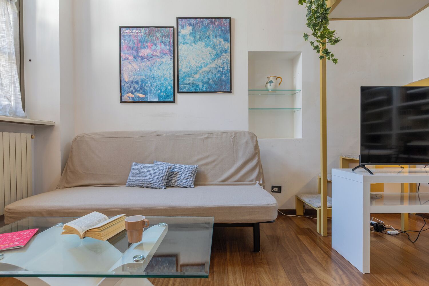 A living room with a beige sofa, two framed artworks, a glass coffee table with books and a mug, a TV on a stand, and a small shelf with a decorative item.