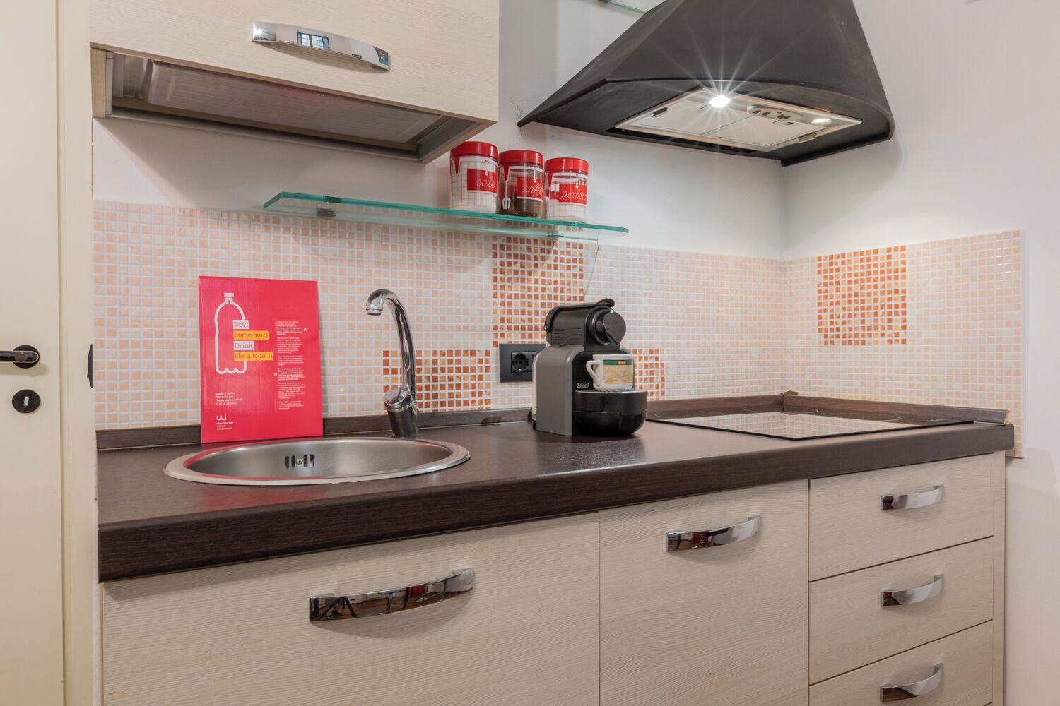 A modern kitchen with a stainless steel sink, a coffee machine, and a glass shelf with jars.