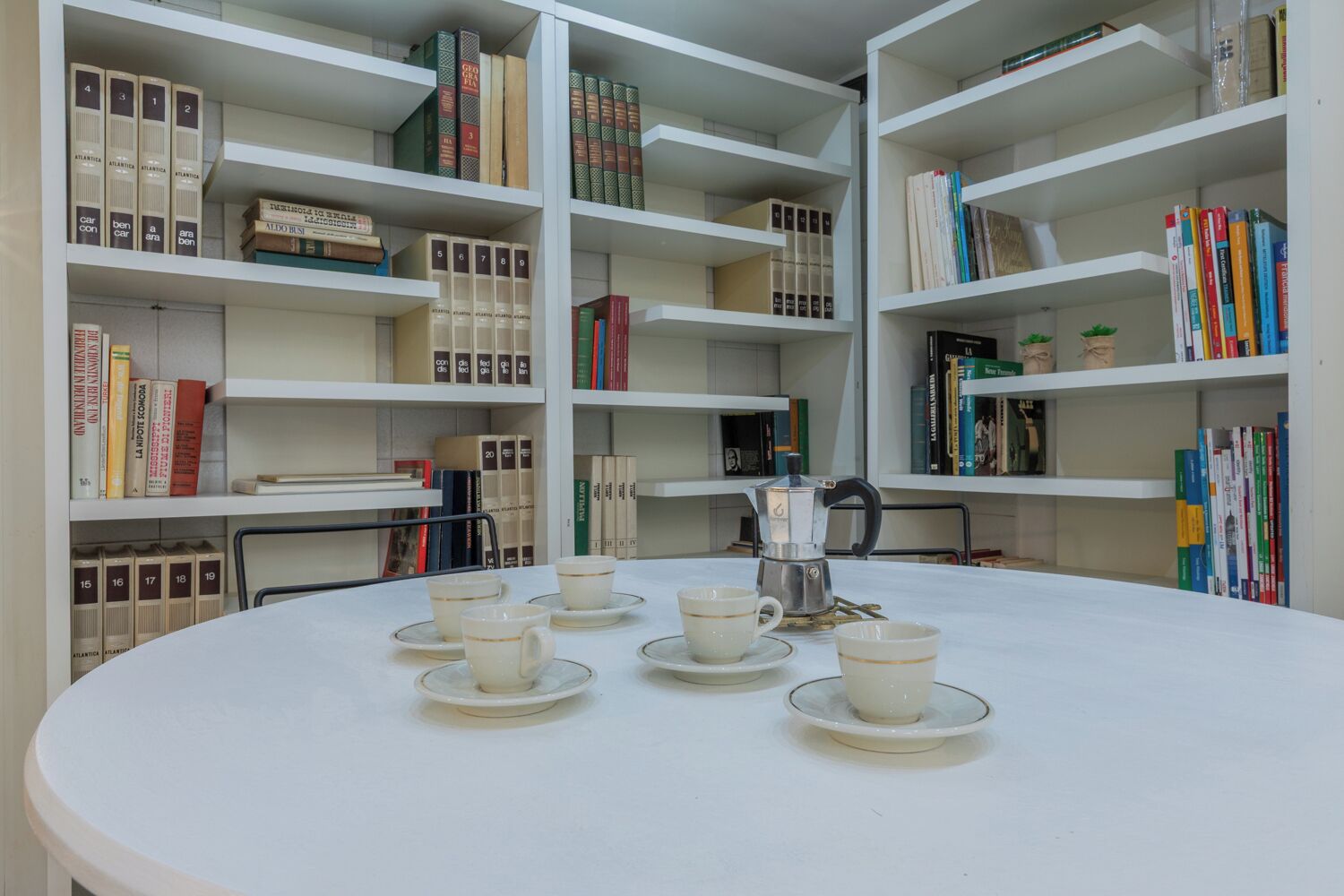 A library with white shelves, books, and a table with cups and a coffee pot.