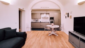 Ein modernes Wohnzimmer mit einem dunklen Sofa, einem Esstisch mit Stühlen, einem Fernseher und einer Küche im Hintergrund.