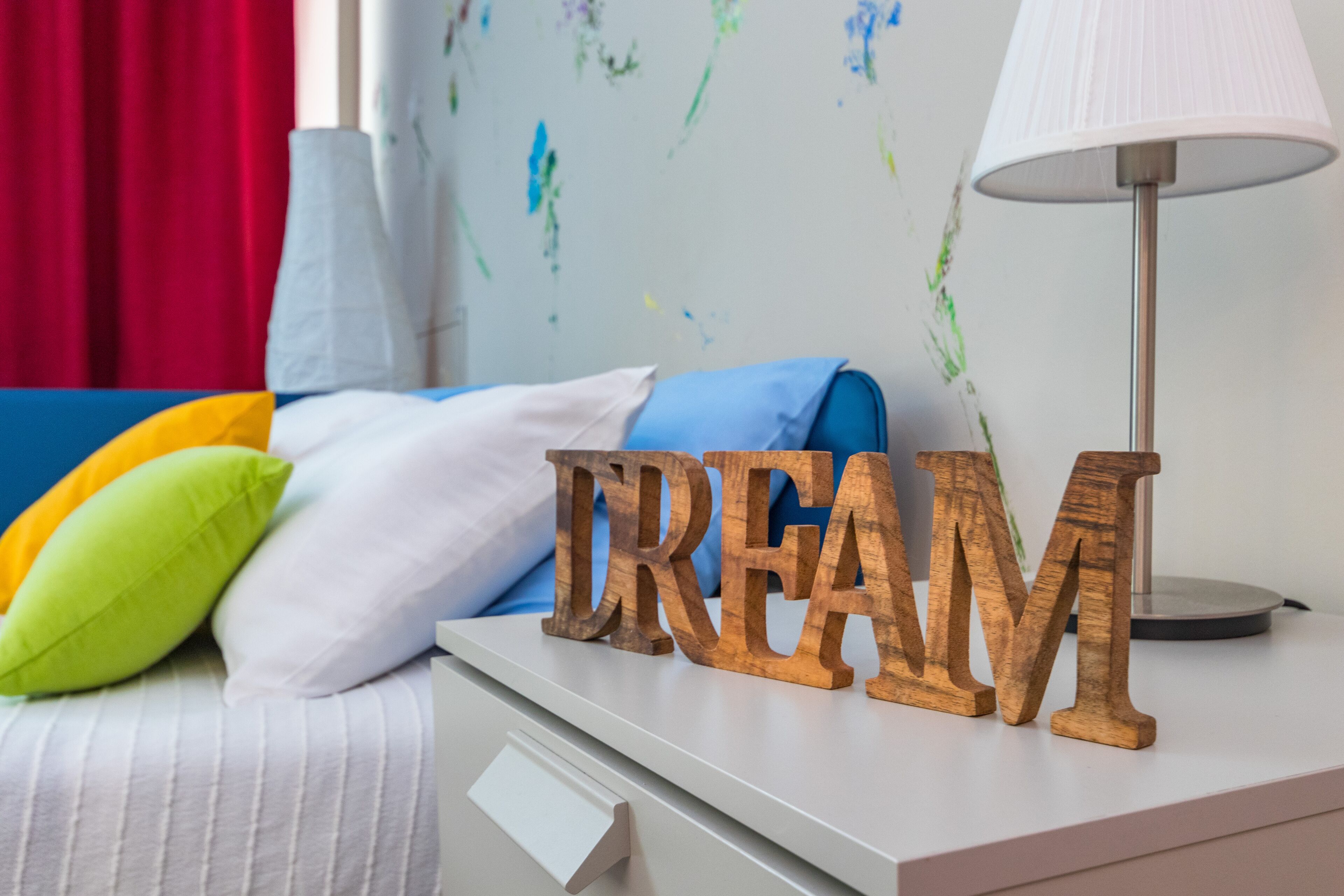 A bed with a white headboard, a blue pillow, and a green pillow. There is a wooden sign with the word "DREAM" on a white nightstand.