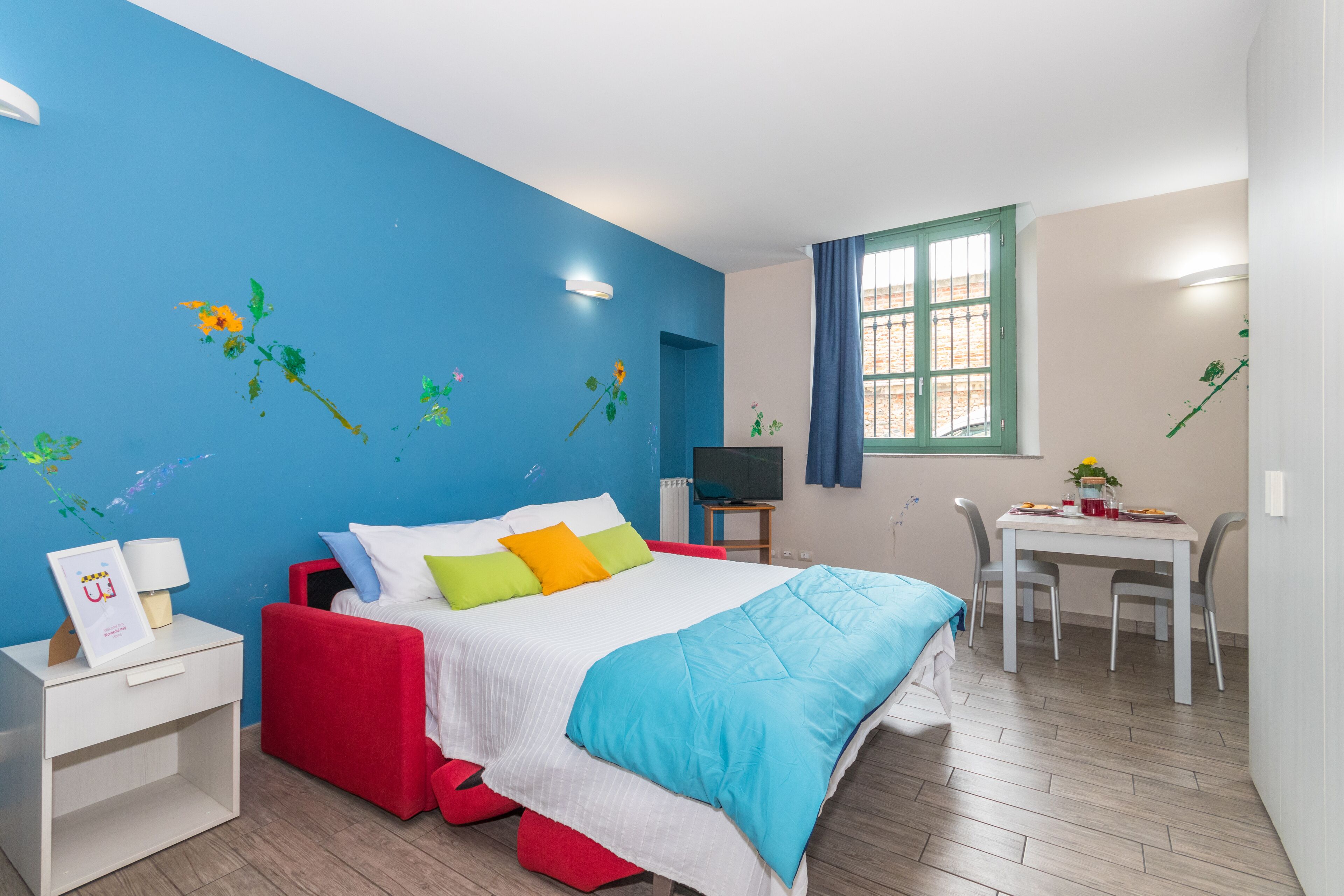 A bedroom with a bed, bedside table, TV, and a dining area with chairs and a table.