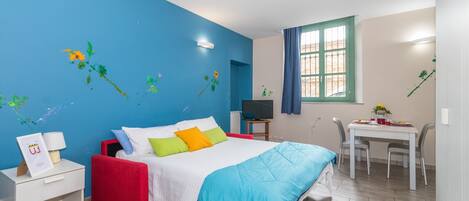A bedroom with a bed, bedside table, TV, and a dining area with chairs and a table.