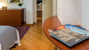 Un dormitorio con un aparador de madera, una cama con manta gris, una alfombra roja estampada y un libro titulado 'Valentino' sobre una mesita de noche de madera.
