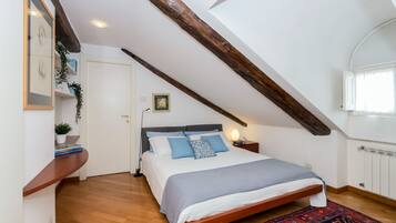 Ein Schlafzimmer mit schrägverdeckter Decke, Holzbalken, einem Bett mit blau-weißer Bettwäsche, einem Nachttisch mit Lampe und einem gemusterten Teppich.