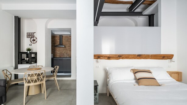 Una habitación moderna con cama de madera y cabecera de madera, una mesa de comedor redonda con sillas, una zona de cocina y un pequeño armario de madera.