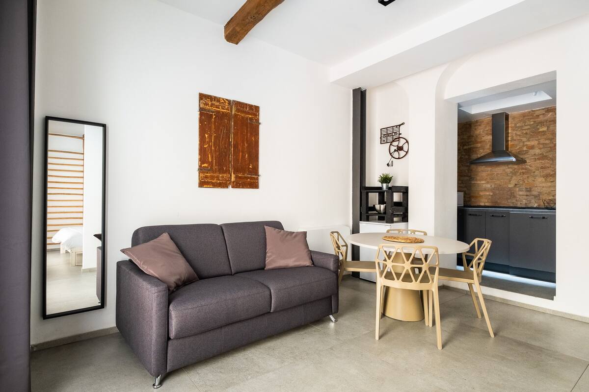 A modern living room with a gray sofa, wooden dining table, and chairs. There is a kitchen visible through an archway with a stone backsplash and a chimney.