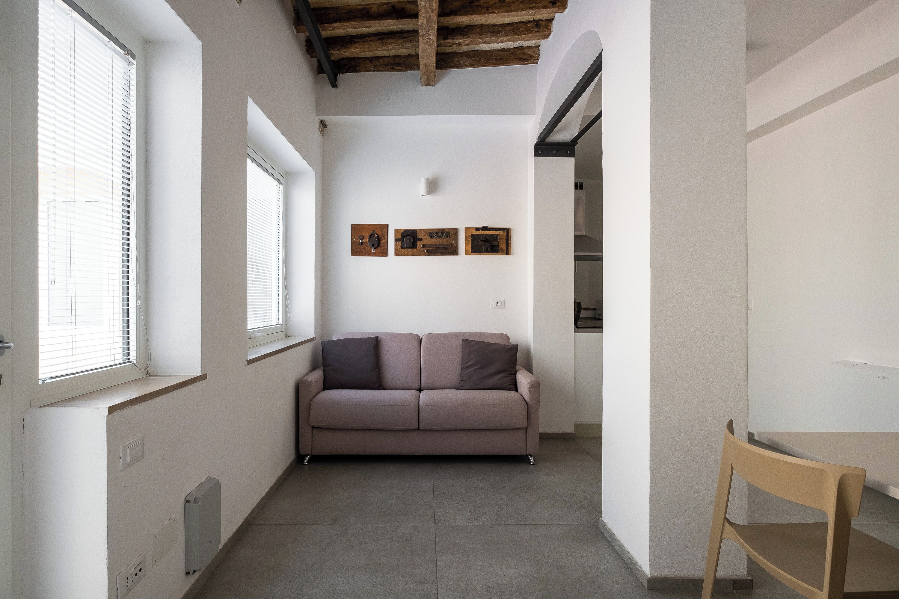 A narrow hallway with a sofa and a chair, white walls, and a wooden ceiling beam.